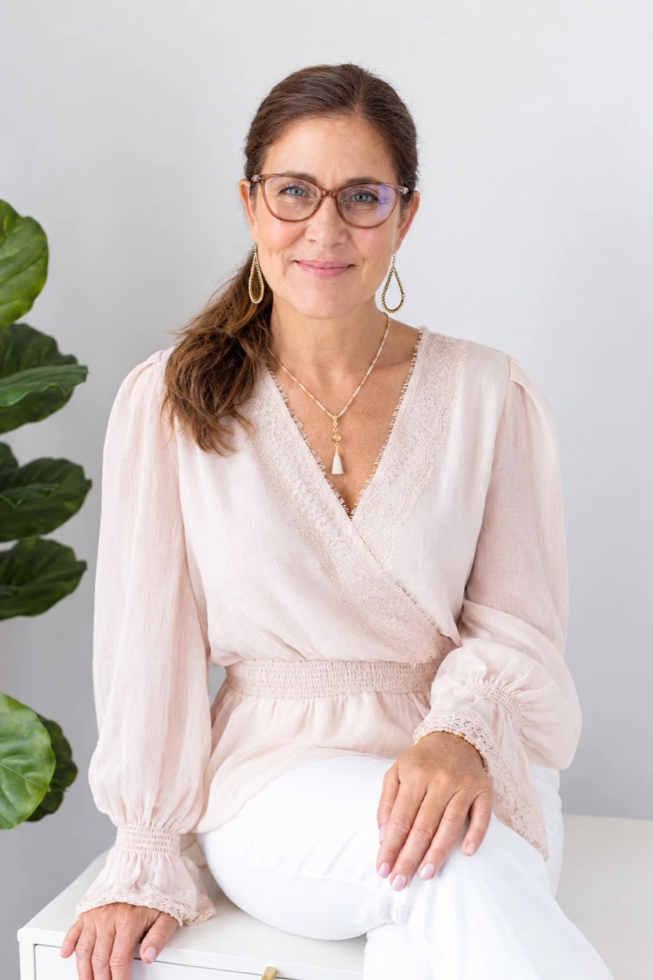 Sienna Smith smiling warmly in a blush top and glasses, seated beside a plant